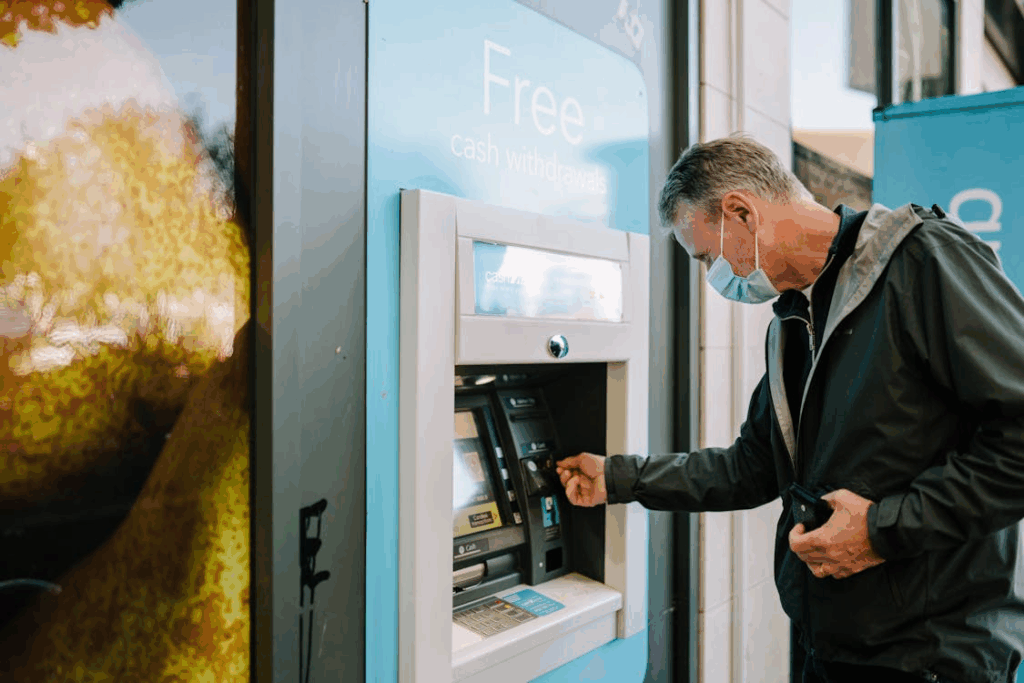 A man wearing a face mask and a dark jacket is using an ATM for free cash withdrawals, inserting or removing his bank card.
